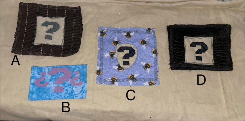 May include: Four fabric patches with question mark designs. One patch is brown with a white square and a blue question mark. Another is blue with a pink question mark. A third is blue with bee patterns and a black question mark. The last is black with a white square and a blue question mark.