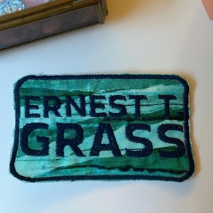 May include: A rectangular embroidered patch featuring a green and teal watercolour-style background with the text ERNEST T. GRASS stitched in bold, dark navy blue thread. The patch has a dark navy blue border and rounded corners.