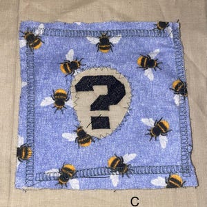 May include: A square, blue fabric patch with a repeating pattern of yellow and black bees with white wings. A beige, question mark applique is centered on the patch. The letter "C" is printed in the bottom right corner.