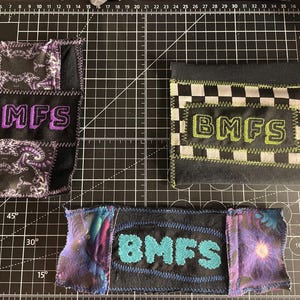 May include: Three fabric patches with the embroidered letters "BMFS". One is black with purple lettering, another black with green lettering, and the third black with teal lettering. Each patch features a unique patterned fabric, all handmade.