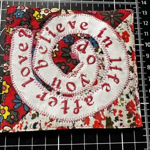 May include: A square fabric patch with floral patterns in red, white, and green. The patch features a white, circular design with the words "believe in life after love" stitched in red thread. The edges are also stitched in red.