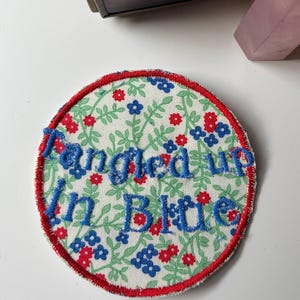 May include: A circular embroidered patch featuring a white background with a repeating pattern of green vines and small red and blue flowers. The text "Tangled up In Blue" is stitched in blue thread across the center, bordered by a red edge.