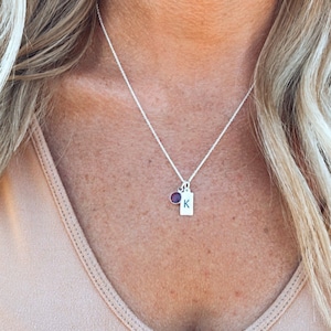 Op de afbeelding: Een zilveren ketting met een kleine zilveren hanger met de letter "K" erop. De hanger is bevestigd aan een kleine paarse edelsteen.