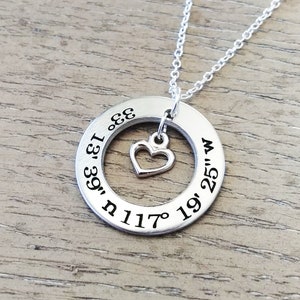 May include: Silver-toned necklace featuring a circular pendant with engraved coordinates and a small heart charm. The coordinates are engraved around the circle. The necklace hangs on a delicate chain, perfect for everyday wear.