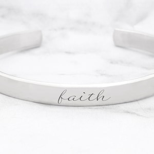 Puede incluir: Un brazalete de plata con la palabra "faith" grabada en la parte delantera.