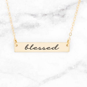 Puede incluir: Un collar de oro con un colgante de barra que dice "blessed" en letra cursiva.