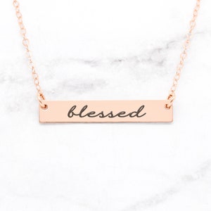 Puede incluir: Un collar de oro rosa con un colgante de barra que dice "blessed" en letra cursiva.