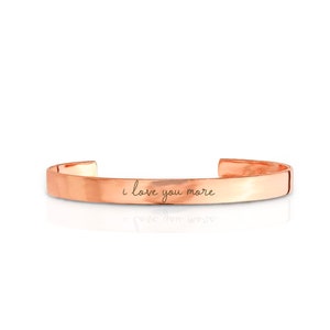 May include: A rose gold cuff bracelet with the inscription "i love you more" engraved on the front.