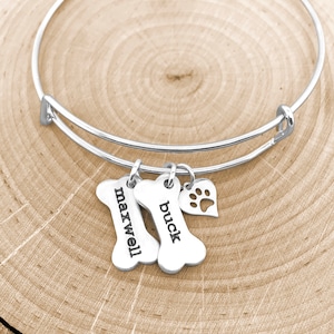 May include: Silver charm bracelet with two bone-shaped charms engraved with the names "Maxwell" and "Buck" and a heart charm with a paw print cutout.