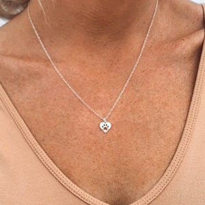 May include: A silver necklace with a heart pendant featuring a paw print cutout. The necklace is worn by a person with a light brown top.