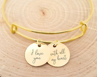 Puede incluir: Pulsera de brazalete de oro con dos dijes redondos. Los dijes tienen el texto "Te amo" y "con todo mi corazón" grabado en ellos.