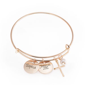 Può includere: Braccialetto rigido in oro con due ciondoli rotondi, uno inciso con "Sophia" e l'altro con "Jeremiah 29:11". Il braccialetto ha anche un piccolo ciondolo a forma di croce e una perla.