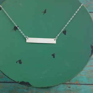 May include: A silver bar necklace with a simple, rectangular pendant hanging from a delicate chain. The necklace is displayed on a green plate.