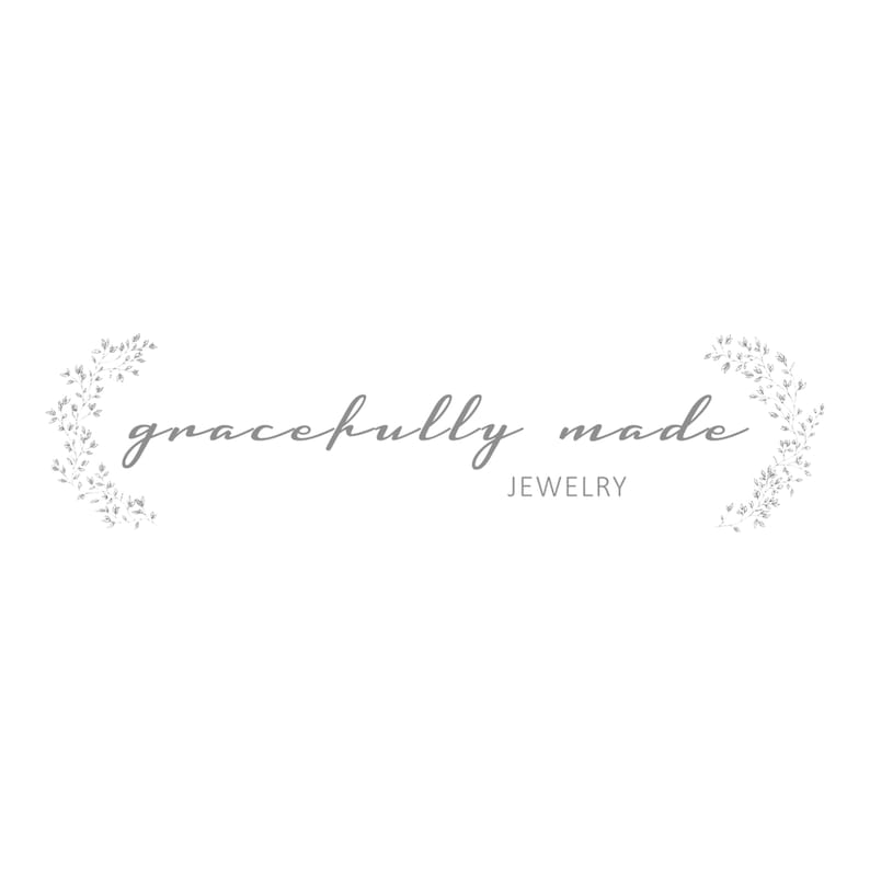 Może przedstawiać: Logo firmy jubilerskiej o nazwie "gracefully made". Logo zawiera nazwę firmy w ozdobnym, pochylonym kroju pisma, z kwiatowym wzorem po obu stronach.