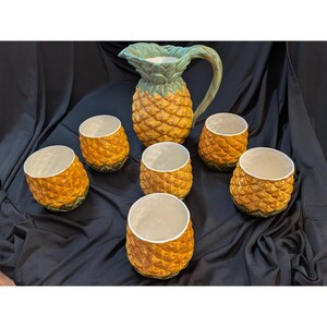 Puede incluir: Una jarra de cerámica con forma de piña con una parte superior y asa de hojas verdes, acompañada de seis vasos a juego con textura de piña. Cada pieza presenta un exterior amarillo anaranjado con un patrón de escamas en relieve y un interior blanco roto.