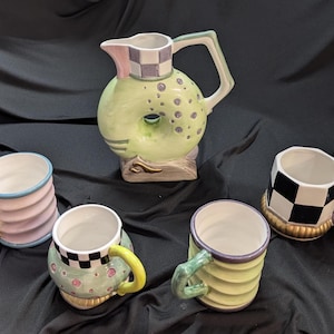 Puede incluir: Una jarra de cerámica con un orificio central, borde a cuadros y puntos verdes, acompañada de cuatro tazas. Las tazas presentan diseños variados que incluyen estrías en tonos pastel, cuadros blancos y negros, y patrones verdes.
