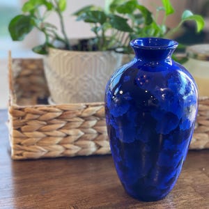 May include: A tall, cobalt blue vase with a glossy finish. The vase has a textured, mottled pattern in shades of dark and light blue. The vase is empty and sits on a wooden surface, with a woven basket and potted plant in the background.