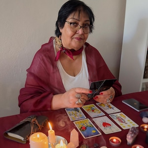 May include: A woman with glasses and a red scarf is holding tarot cards. Several tarot cards are laid out on a red table, along with lit candles, a leather-bound journal, and crystals. The scene suggests a reading or divination session.