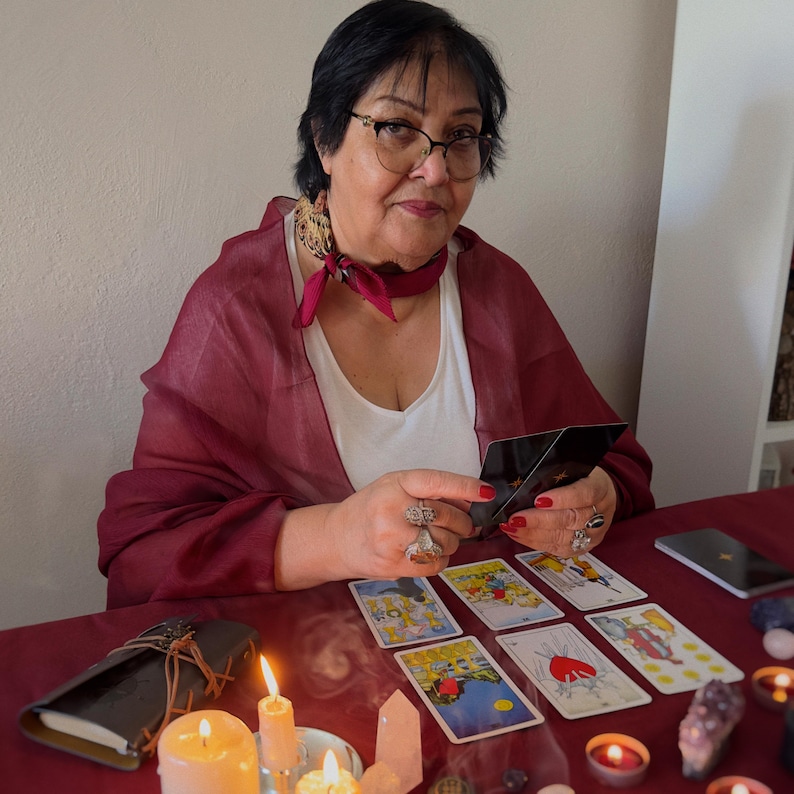 May include: A woman with glasses and a red scarf is holding tarot cards. Several tarot cards are laid out on a red table, along with lit candles, a leather-bound journal, and crystals. The scene suggests a reading or divination session.