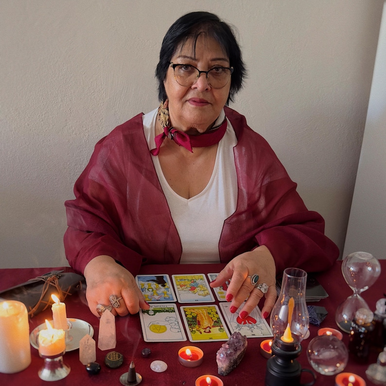 May include: A woman with glasses sits at a table with tarot cards, candles, and crystals. She wears a white top, a burgundy scarf, and a matching shawl. The table is covered with a red cloth, and there are lit candles and a small oil lamp.