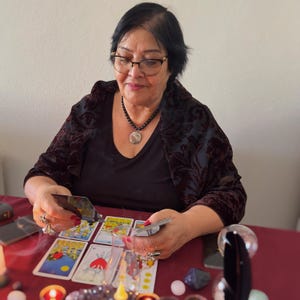 May include: A person with dark hair and glasses wearing a black top and dark velvet shawl, holding tarot cards over a red surface. Several tarot cards are laid out, including the Three of Swords, alongside small crystals and lit candles.