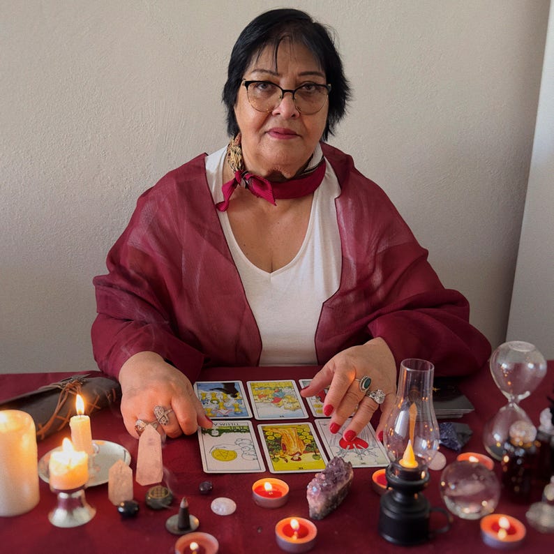 May include: A woman with glasses sits at a red table, surrounded by candles, crystals, and tarot cards. She wears a white top, a burgundy shawl, and a red scarf. The scene evokes a mystical or spiritual theme.