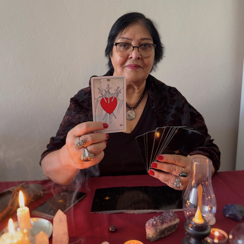 May include: A woman holding a tarot card with a red heart pierced by three swords. She is surrounded by candles, crystals, and other tarot cards. The card is from a tarot deck.