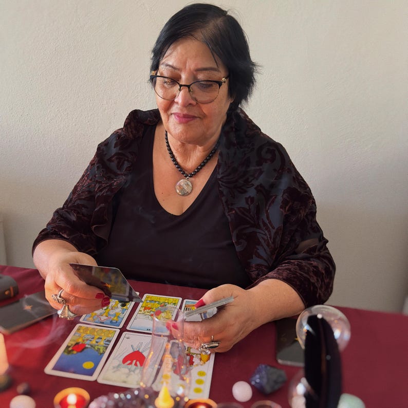 May include: A person with dark hair and glasses wearing a black top and dark velvet shawl, holding tarot cards over a red surface. Several tarot cards are laid out, including the Three of Swords, alongside small crystals and lit candles.