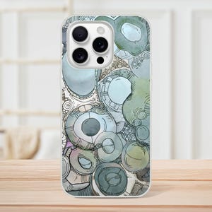 Könnte beinhalten: Eine transparente Handyhülle mit einem Aquarelldesign aus sich überlappenden Kreisen in Blau-, Grün- und Grautönen. Die Hülle hat eine glänzende Oberfläche und ein schützendes Design mit Aussparungen für Kamera und Tasten des Telefons. Das Design umfasst komplizierte Liniendetails.