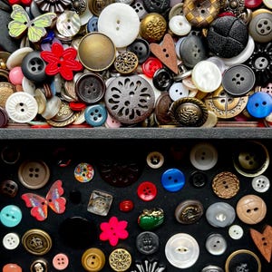 May include: A collection of various buttons in a black box. The buttons are in different sizes, shapes, and colors, including red, white, blue, and gold. Some buttons have floral and butterfly designs.