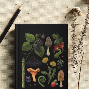 May include: A black notebook featuring botanical illustrations of various plants and fungi, including blackberries, dandelions, and mushrooms. A black pencil rests on the beige fabric surface next to the notebook.