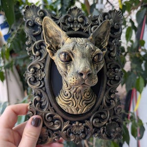 Könnte beinhalten: Eine dreidimensionale Wandplakette mit einem skulptierten Sphynx-Katzenkopf in metallischer Bronzeoptik, eingefasst in einen verzierten, rechteckigen schwarz-bronzenen Rahmen mit komplexen Schnörkeln und floralen Details um das ovale Zentrum.