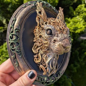 Peut inclure: Une plaque murale en trois dimensions représentant une tête d'écureuil sculptée en or et bronze, montée sur une base ovale noire. Le cadre est orné de motifs en filigrane verts et bronze, créant un aspect texturé et superposé.