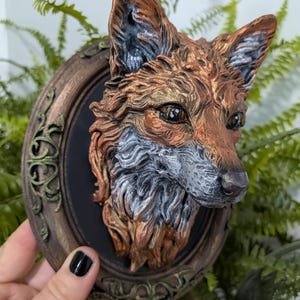 Può includere: Una placca da parete tridimensionale raffigurante una testa di volpe scolpita con riflessi metallici color rame, bronzo e argento. La volpe è montata su una cornice ovale scura con dettagli ornamentali a forma di vite verde invecchiata.