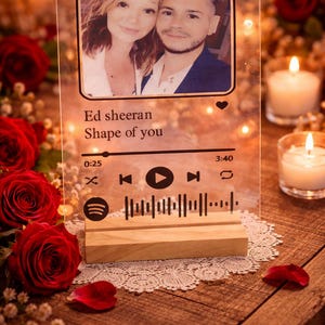 Può includere: Un espositore fotografico in acrilico trasparente con la foto di una coppia, il titolo della canzone "Shape of You" di Ed Sheeran e una barra musicale in stile Spotify. Poggia su una base di legno, circondato da rose rosse e candele.