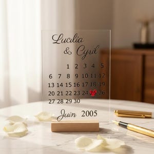Puede incluir: Calendario de boda de acrílico transparente con los nombres "Lucilia & Cyril" y la fecha "Junio 2005". El 25 de junio está resaltado en rojo. El calendario está sobre una base de madera.