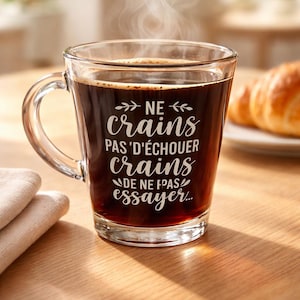 Peut inclure: Une tasse en verre transparente remplie de café noir, de la vapeur s'élevant du dessus. La tasse a une anse et présente un texte blanc qui dit "NE crains pas d'échouer, crains de ne pas essayer..." Un croissant est posé sur une assiette en arrière-plan.