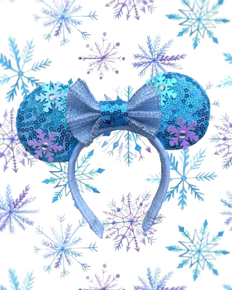 May include: A light blue headband with sequined ears and a matching bow. The ears are decorated with light blue sequins and purple snowflake accents. The background features a pattern of blue and purple snowflakes.