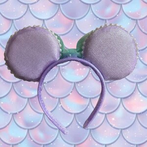 May include: A lavender-colored headband with two round, padded ears. The ears are trimmed with white beads and have a shimmering texture. A light green bow sits between the ears. The background features a mermaid scale pattern in shades of purple, pink, and blue.