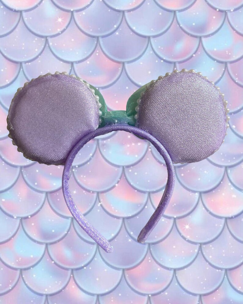 May include: A lavender-colored headband with two round, padded ears. The ears are trimmed with white beads and have a shimmering texture. A light green bow sits between the ears. The background features a mermaid scale pattern in shades of purple, pink, and blue.