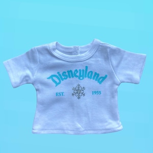 Puede incluir: Camiseta blanca con la palabra "Disneyland" en escritura azul claro. Debajo, un copo de nieve plateado y el texto "EST. 1955". La camiseta tiene mangas cortas y cuello redondo.