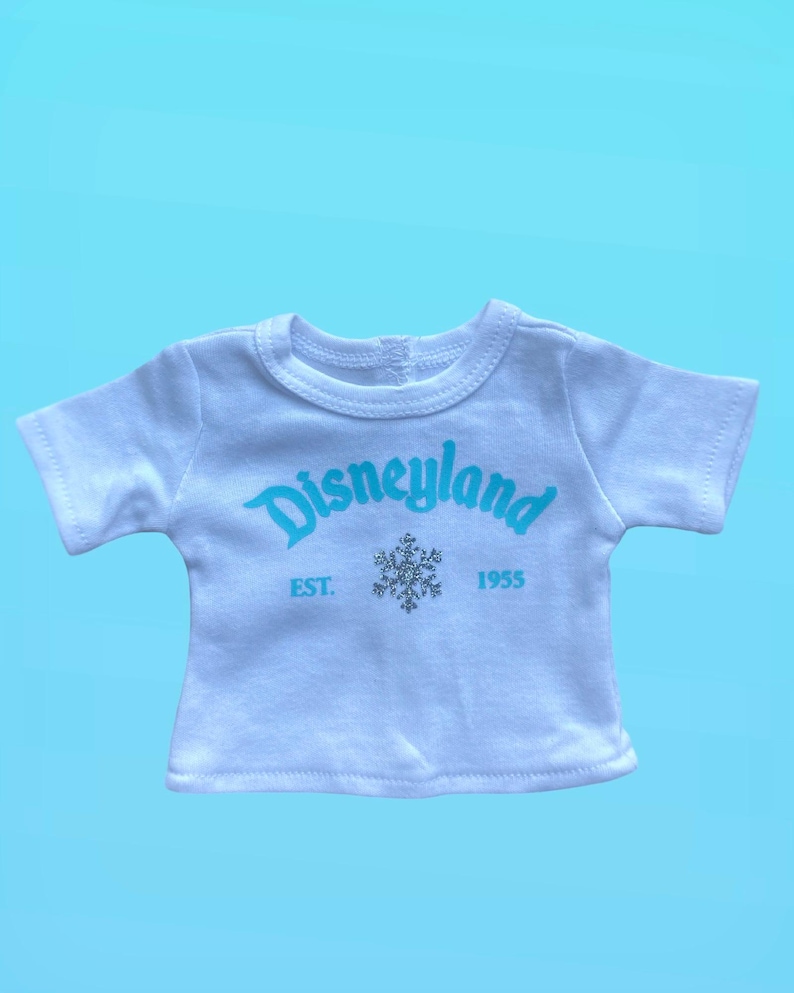 Puede incluir: Camiseta blanca con la palabra "Disneyland" en escritura azul claro. Debajo, un copo de nieve plateado y el texto "EST. 1955". La camiseta tiene mangas cortas y cuello redondo.