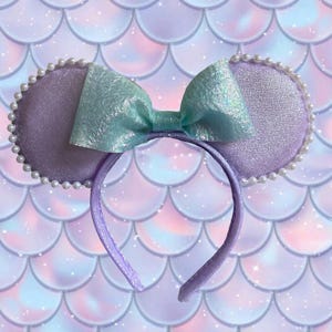 May include: A purple headband with two round, lavender ears trimmed with white pearls. A large, iridescent, light blue bow sits between the ears. The background is a mermaid scale pattern in shades of purple, pink, and blue.