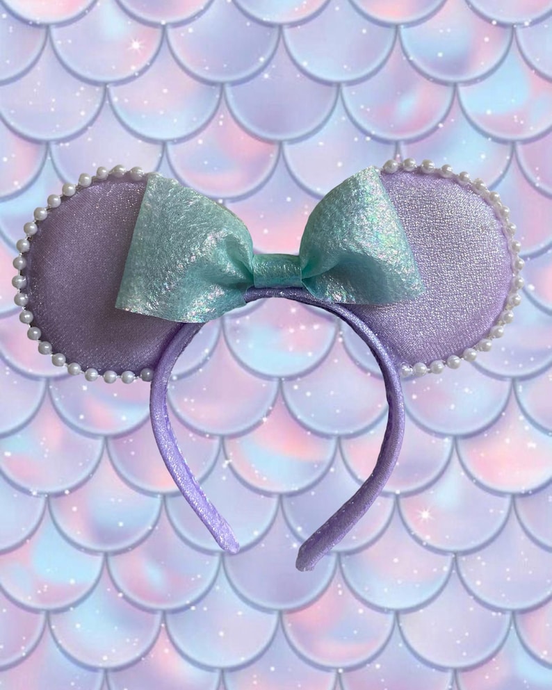 May include: A purple headband with two round, lavender ears trimmed with white pearls. A large, iridescent, light blue bow sits between the ears. The background is a mermaid scale pattern in shades of purple, pink, and blue.
