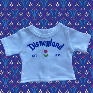 Op de afbeelding: Een lichtblauw T-shirt met het woord "Disneyland" in blauw script, een rode tulpafbeelding en de tekst "EST 1955". Het shirt ligt op een blauwe achtergrond met patronen.