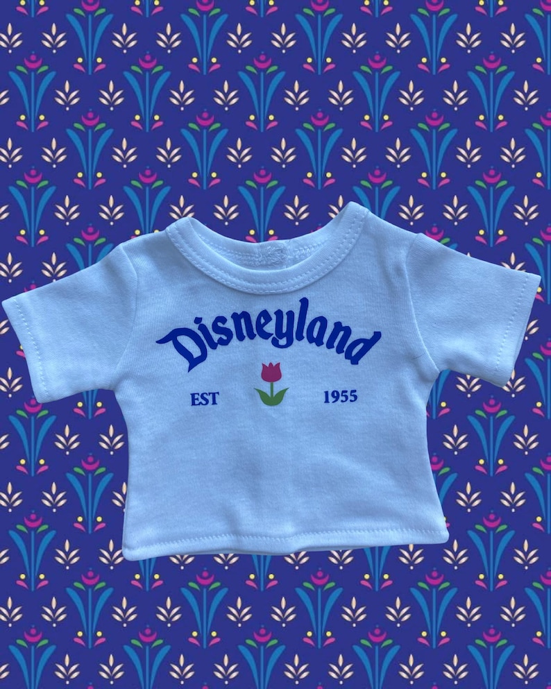 Op de afbeelding: Een lichtblauw T-shirt met het woord "Disneyland" in blauw script, een rode tulpafbeelding en de tekst "EST 1955". Het shirt ligt op een blauwe achtergrond met patronen.