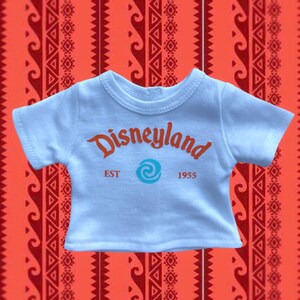 K&ouml;nnte beinhalten: Hellblaues T-Shirt mit dem Wort "Disneyland" in orangefarbener Schrift, einem t&uuml;rkisfarbenen Wirbel und dem Text "EST 1955". Das Shirt befindet sich vor einem roten Hintergrundmuster.