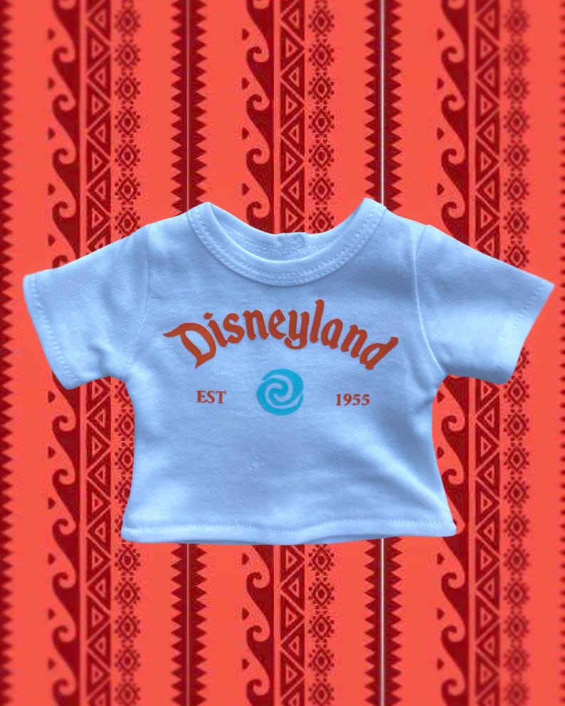 K&ouml;nnte beinhalten: Hellblaues T-Shirt mit dem Wort "Disneyland" in orangefarbener Schrift, einem t&uuml;rkisfarbenen Wirbel und dem Text "EST 1955". Das Shirt befindet sich vor einem roten Hintergrundmuster.
