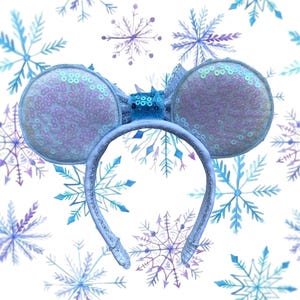 May include: A light blue headband with sequined mouse ears. The ears are round and covered in iridescent sequins. A small bow and decorative accents sit between the ears. The background is white with blue and purple snowflake designs.