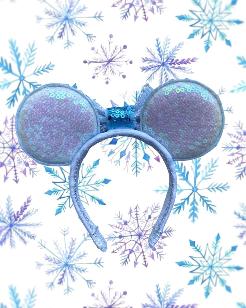 May include: A light blue headband with sequined mouse ears. The ears are round and covered in iridescent sequins. A small bow and decorative accents sit between the ears. The background is white with blue and purple snowflake designs.
